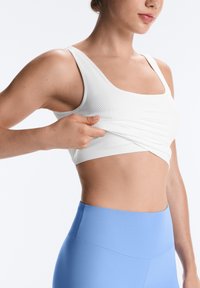 Femme ajustant un soutien-gorge de sport blanc, portant des leggings bleu clair taille haute, montrée du milieu du torse jusqu'au menton sur un fond uni.