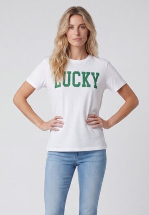 Kobieta o długich blond włosach, ubrana w białą koszulkę z napisem "LUCKY" zielonymi literami i jasnoniebieskie dżinsy, stoi z rękami na biodrach.