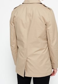 Cappotto trench beige realizzato in tessuto leggero, con una fessura sul retro, spalline sulle spalle e un design di colletto classico.