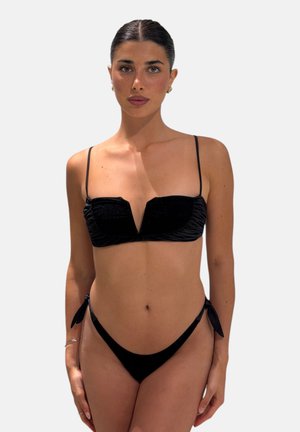 Donna con capelli scuri raccolti in uno chignon ordinato, che indossa un bikini nero con sottili spalline e scollatura a V, in piedi davanti a uno sfondo semplice grigio chiaro.