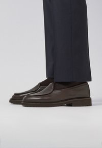 Bruine leren instapschoenen met een lichte glans, ronde neus en gestructureerd oppervlak, gecombineerd met donkere broek, weergegeven tegen een neutrale achtergrond.