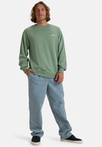 Hellgrüner Pullover mit gerippten Bündchen und Saum, mit weißem Logo-Text; getragen mit lockeren, hellblauen Jeans und dunklen Turnschuhen.