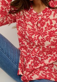 Blusa floreale rossa e crema con bottoni, realizzata in un tessuto leggero e testurizzato, e con scollatura rotonda. Abbinata a jeans in denim blu.