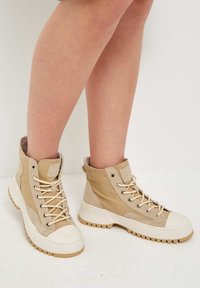 Nohy obuté v béžových vysokých teniskách s tlustou bílou podrážkou, tkaničkami a drsným vzorkem, stojící na světlé podlaze proti bílé zdi.