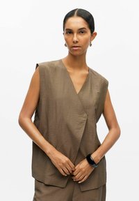 Top senza maniche marrone con scollatura a V, texture morbida e design anteriore sovrapposto. Indossato con un bottom coordinato e un bracciale nero.