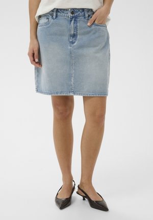 Femeie purtând fustă din denim bleu deschis, top alb și pantofi negri cu toc mic, cu bretea și vârf ascuțit, stând cu o mână în buzunar.