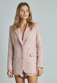 Blazer rosa con paillettes e un tessuto a trama, con revers a punta, due tasche frontali e bottoni decorativi. Indossato sopra dei pantaloni corti coordinati.