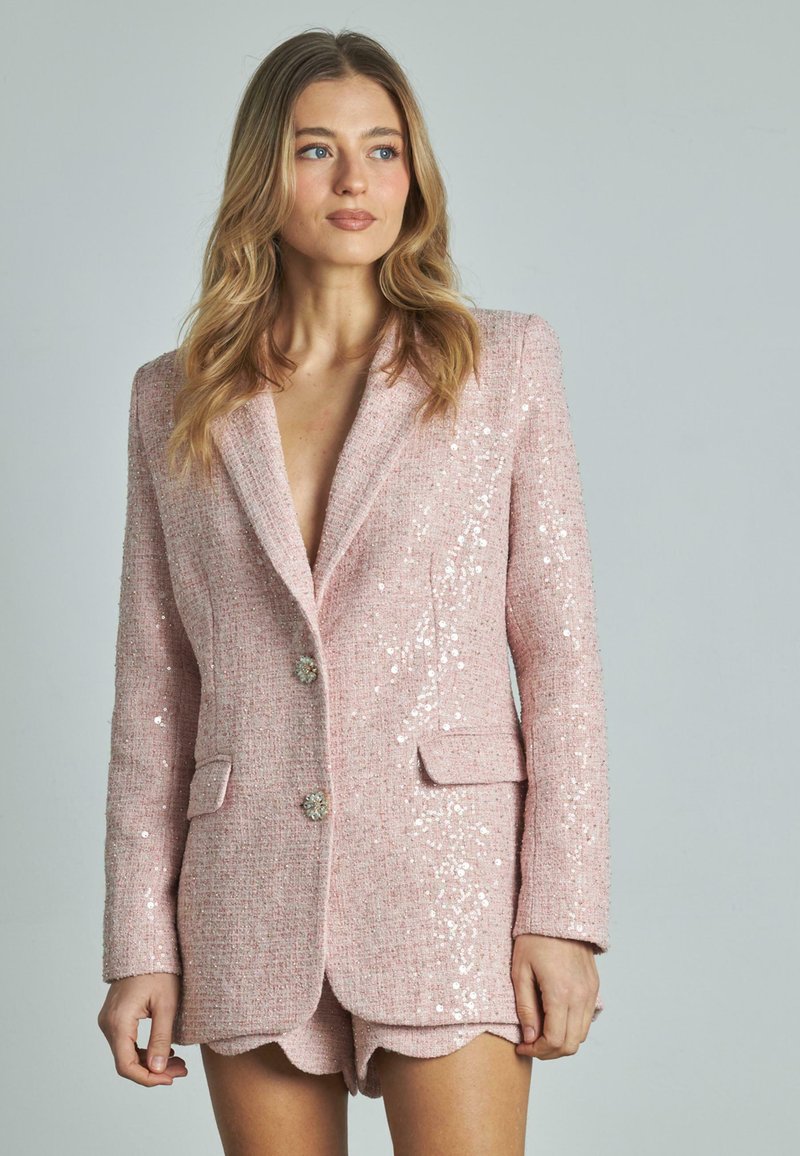 Blazer rosa con paillettes e un tessuto a trama, con revers a punta, due tasche frontali e bottoni decorativi. Indossato sopra dei pantaloni corti coordinati.