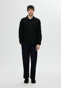 Juodas marškinėliai su sagomis ir dviem kišenėmis priekyje, derinami su laisvais tamsiai mėlynais kelnais ir juodais batais, prieš paprastą foną.