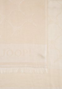 Textile tissé beige présentant un motif floral, avec des bords fringés et le mot "JOOP!" subtilement intégré dans le design.