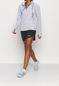 Maglione grigio a maniche lunghe con zip a quarto, abbinato a pantaloni corti neri con spacchi laterali. Scarpe da corsa azzurro chiaro con dettagli bianchi.