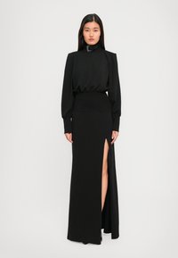 Eine Frau trägt ein langes schwarzes Kleid mit vollem Ärmeln, einem hohen Hals mit Schnalle und einem schulterhohen Schlitz, während sie vor einem schlichten hellen Hintergrund steht.