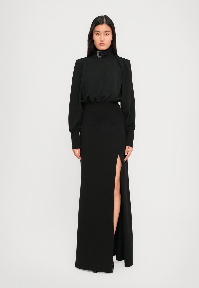 Eine Frau trägt ein langes schwarzes Kleid mit vollem Ärmeln, einem hohen Hals mit Schnalle und einem schulterhohen Schlitz, während sie vor einem schlichten hellen Hintergrund steht.