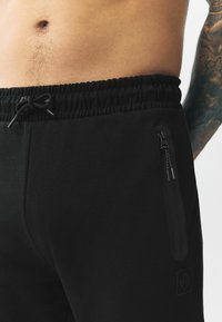 Schwarze Jogginghose mit elastischem Bund und Kordelzug. Verfügt über eine Reißverschlusstasche an der Seite und ein dezentes Logo-Detail. Glatte Textur.