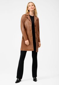 Brun læderjakke med skræddersyet design, notched krave, tre knapper og sidelommer, båret over en sort top og bukser.