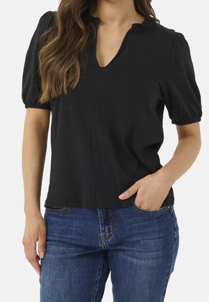 Kobieta ubrana w czarną bluzkę z krótkim rękawem z bufkami i niebieskie jeansy, z jedną ręką w kieszeni, kadrowana na wysokości ramion i bioder.