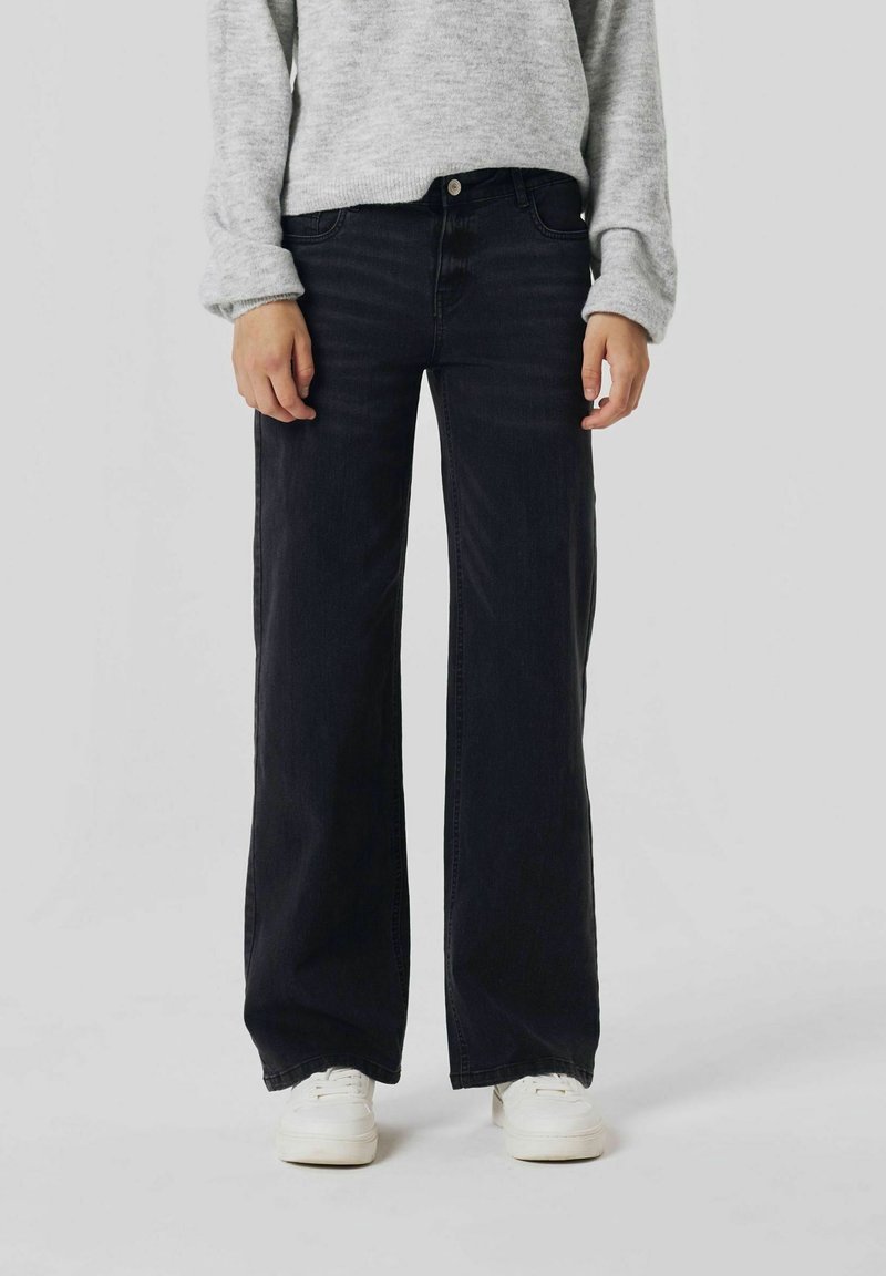 Personne portant un jean noir à jambes larges, un pull gris clair et des baskets blanches, posant devant un fond uni clair.