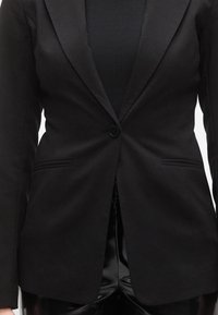 Μαύρο προσαρμοσμένο σακάκι με ένα κουμπί κλεισίματος, γιακά με πέτο και δύο μπροστινές τσέπες. Φορεμένο πά νω από μαύρο ριμπ ύφασμα τοπ, με λεία υφή.