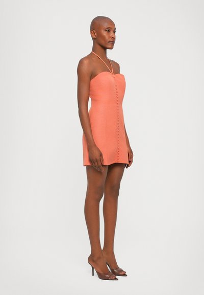 Strapless minidress in koraalkleurige stof met een getailleerd silhouet, voorzien van een sluiting met knopen aan de voorkant en een zachte textuur. Gecombineerd met open teen hakken.