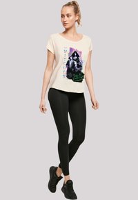 Cremfarbendes T-Shirt mit einem Grafikdesign von "The Witcher." Kurze Ärmel, lockere Passform, dazu schwarze Leggings und schwarze Schuhe.