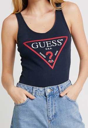 Marineblauwe geribbelde tanktop met een rood driehoeklogo met de tekst "GUESS U.S.A." en een vraagteken; gecombineerd met high-waisted denim shorts.