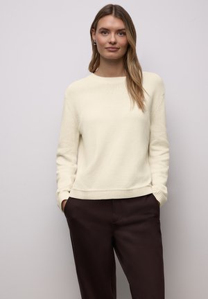 Pull en maille côtelée crème avec un col rond, des manches longues et une coupe décontractée, associé à un pantalon marron foncé.