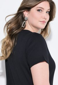 Top nero a maniche corte con effetto rigato e texture, accompagnato da grandi orecchini a goccia scintillanti con forme circolari e rettangolari.