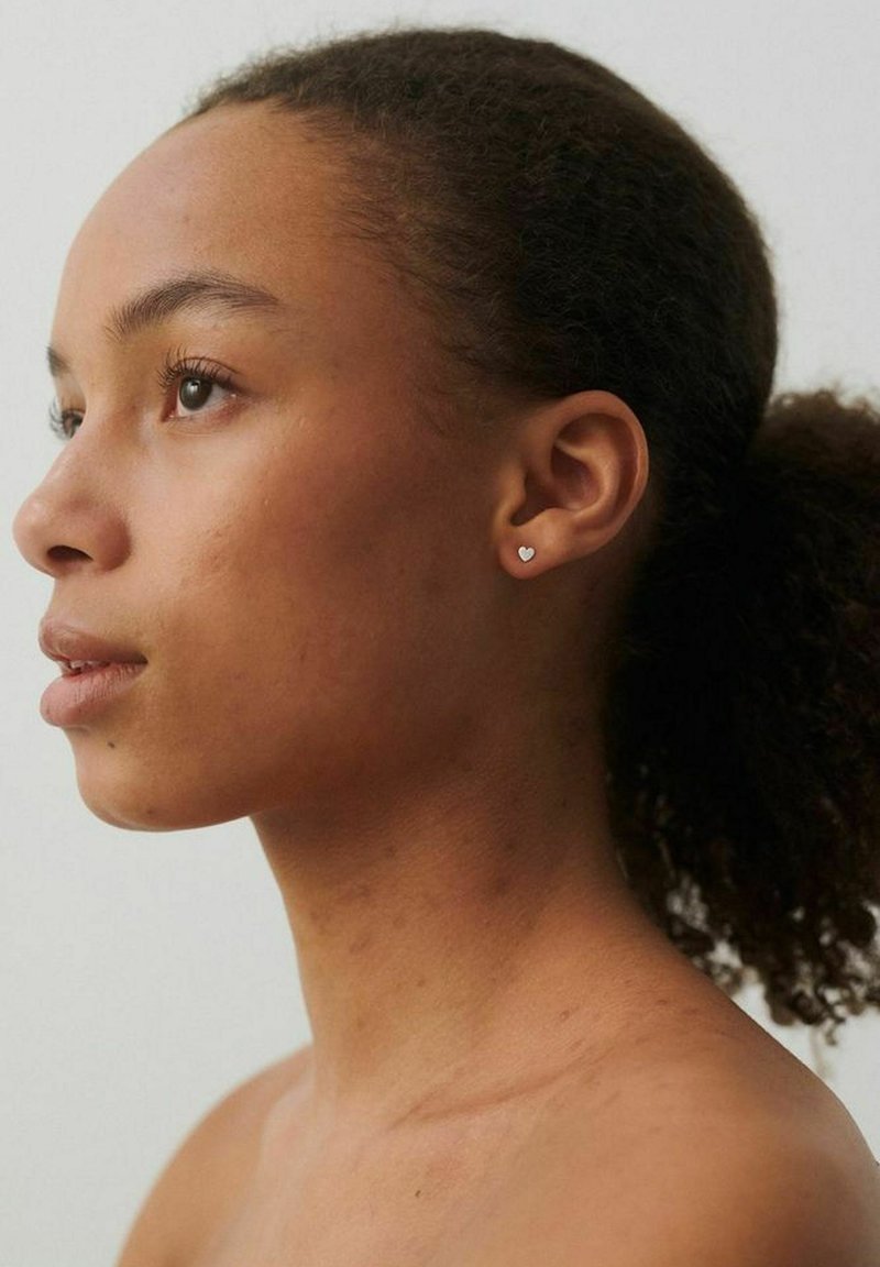 Jeune femme aux cheveux naturels attachés en arrière, portant de petites boucles d'oreilles en forme de cœur, regardant légèrement vers le haut à gauche sur un fond uni.