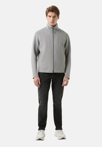 Grijze jas met ritssluiting en een hoge kraag, gemaakt van zacht materiaal. Voorzien van steekzakken en gedragen over een donkere spijkerbroek en witte sneakers.