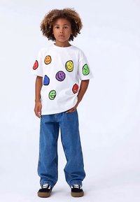 Bērns ar cirtainiem matiem, kas valkā baltu T-kreklu ar krāsainām smaida sejām, zilu džinsu bikses un melnus sporta apavus, stāvot uz baltas fona.