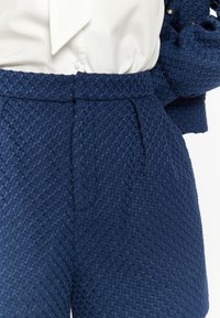Pantaloni corti blu marino, testurizzati, con un motivo a diamante, presentano una vita aderente e delle pieghe. Abbinati a una blusa di colore chiaro.