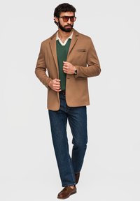 Homme portant un blazer marron sur un pull vert et une chemise blanche, jean bleu, mocassins marron et lunettes de soleil sur fond clair.