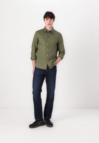 Zöld gombos ing, felgombolt ujjal, farmernadrággal és fekete sneakerrel. Az ing mellén egy kis logó található.