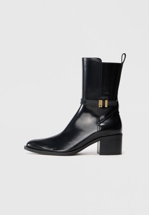 Black leather ankle boot with a mid-height block heel, side strap with gold-tone buckle, and pull tab at the back.