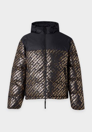 Junger Mann trägt eine schwarze gesteppte Kapuzenjacke mit goldenem Aufdruck, schwarzes Hemd, beige Hose und schwarze Schuhe, steht vor weißem Hintergrund.