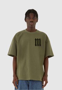 Oliven grøn oversized t-shirt lavet af bomuld; har et sort bogstav "m" logo på venstre brystområde. Blød tekstur, korte ærmer.