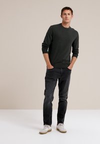 Donkergroene trui met geribbelde manchetten, gecombineerd met donkere jeans en witte sneakers. Model staat tegen een neutrale achtergrond.