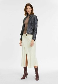 Zwarte leren bikerjack, aansluitende lichtbruine top, vloeiende off-white midi-jurk met een split, en kniehoge bordeauxrode laarzen.