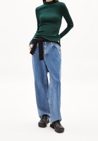 Maglietta a maniche lunghe verde a coste, abbinata a jeans a gamba larga blu chiaro con una cintura nera. Il modello indossa stivali neri con tacco grosso.