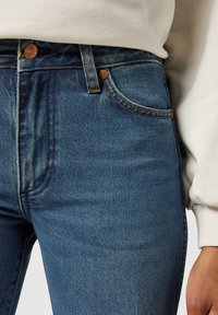 High-waisted blue denim jeans with a faded texture, copper button accents, and subtle stitching details.