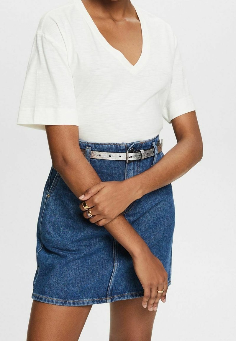 Personne portant un haut blanc à manches courtes et col en V, rentré dans une jupe en jean bleue avec une ceinture argentée, les bras croisés.