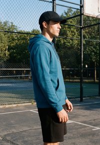 Jeune homme portant un sweat à capuche bleu et une casquette noire, debout sur un terrain de basket extérieur avec une clôture en grillage et des arbres en arrière-plan.