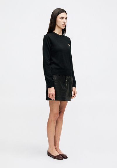 Γυναίκα που στέκεται, φορώντας μαύρο πουλόβερ με μακριά μανίκια και μικρό λογότυπο, μαύρη δερμάτινη φούστα με κουμπιά και καφέ επίπεδα παπούτσια, μπροστά σε απλό φόντο.