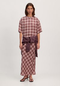 Korte mouw, top met een ruitpatroon in lichtroze en rood, gecombineerd met een langere rok met hetzelfde ruitpatroon. Geaccentueerd met een paarse sweatshirt die om de taille is gebonden.