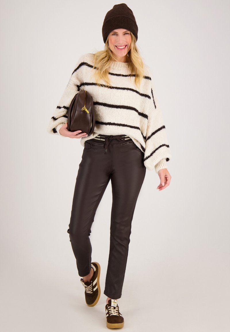 Trui met crèmekleurige en zwarte strepen, bruine nepleren broek, bruine sneakers met crèmekleurige veters, bruine handtas. Model poseert tegen een effen achtergrond.