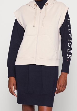 Beżowa bezrękawna bluza z kapturem na suwak na czarnej, długorękawowej sukience z napisem „HILFIGER” na prawym rękawie.