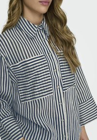 Camisa de botonera a rayas en azul y blanco, con dos bolsillos en el pecho, cuello con solapa y mangas tres cuartos de tejido texturizado.