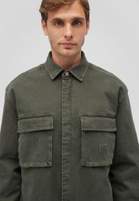 Olivgrüne Denim-Hemdjacke mit Knopfverschluss, zwei Brusttaschen und subtiler Struktur. Sie verfügt über einen Kragen und eine taillierte Passform.