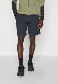 Zwarte shorts met zakken, gecombineerd met zwarte schoenen en sokken. De bovenkant is een lichtgroene jas met donkere mouwen. Gladde stoftextuur.