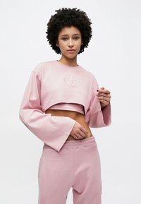 Sweatshirt rosa recortada com mangas sino e um logótipo texturizado na frente, combinada com calças de fato de treino rosa de cintura alta correspondentes. Textura do tecido suave.